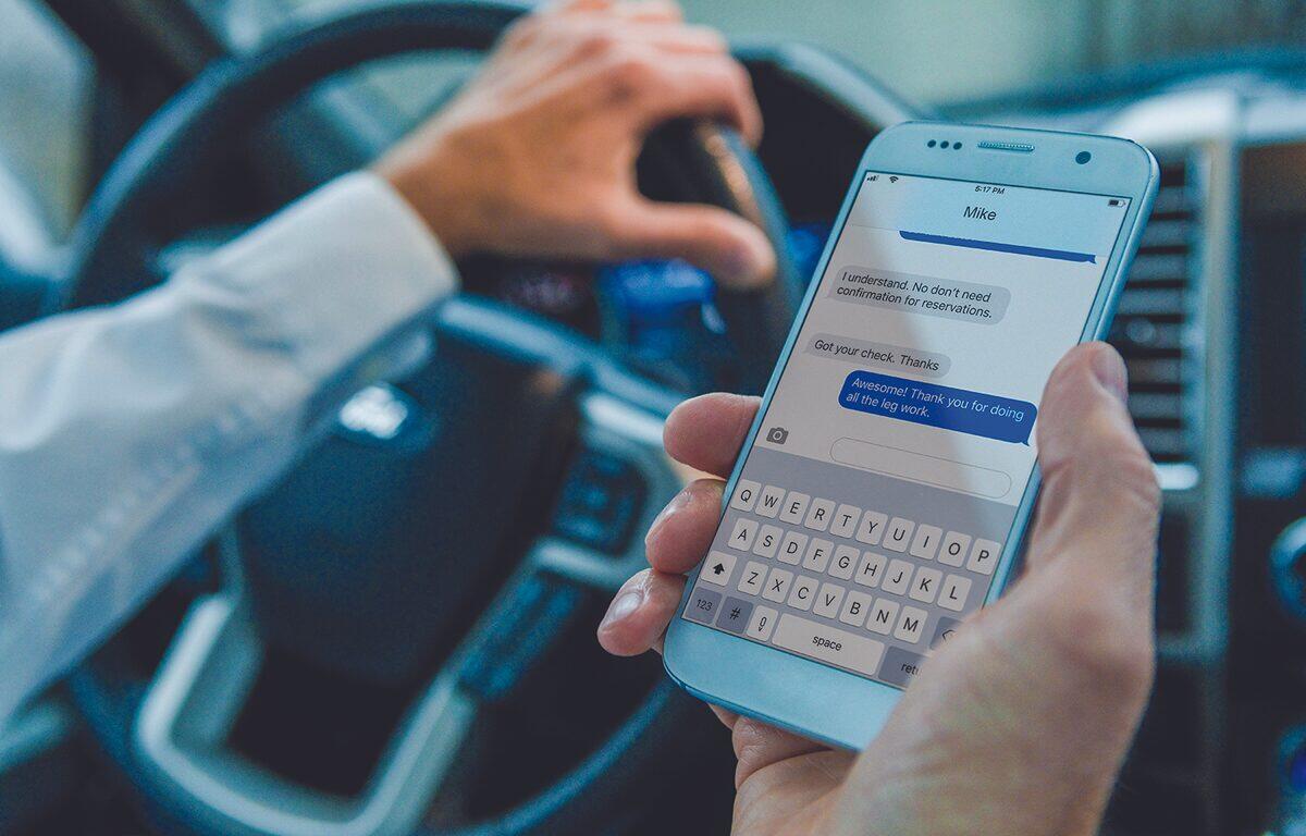 man holds cell phone and steering wheel while driving