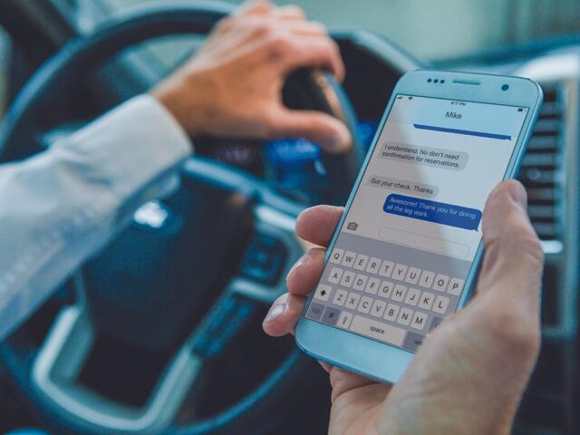 man holds cell phone and steering wheel while driving