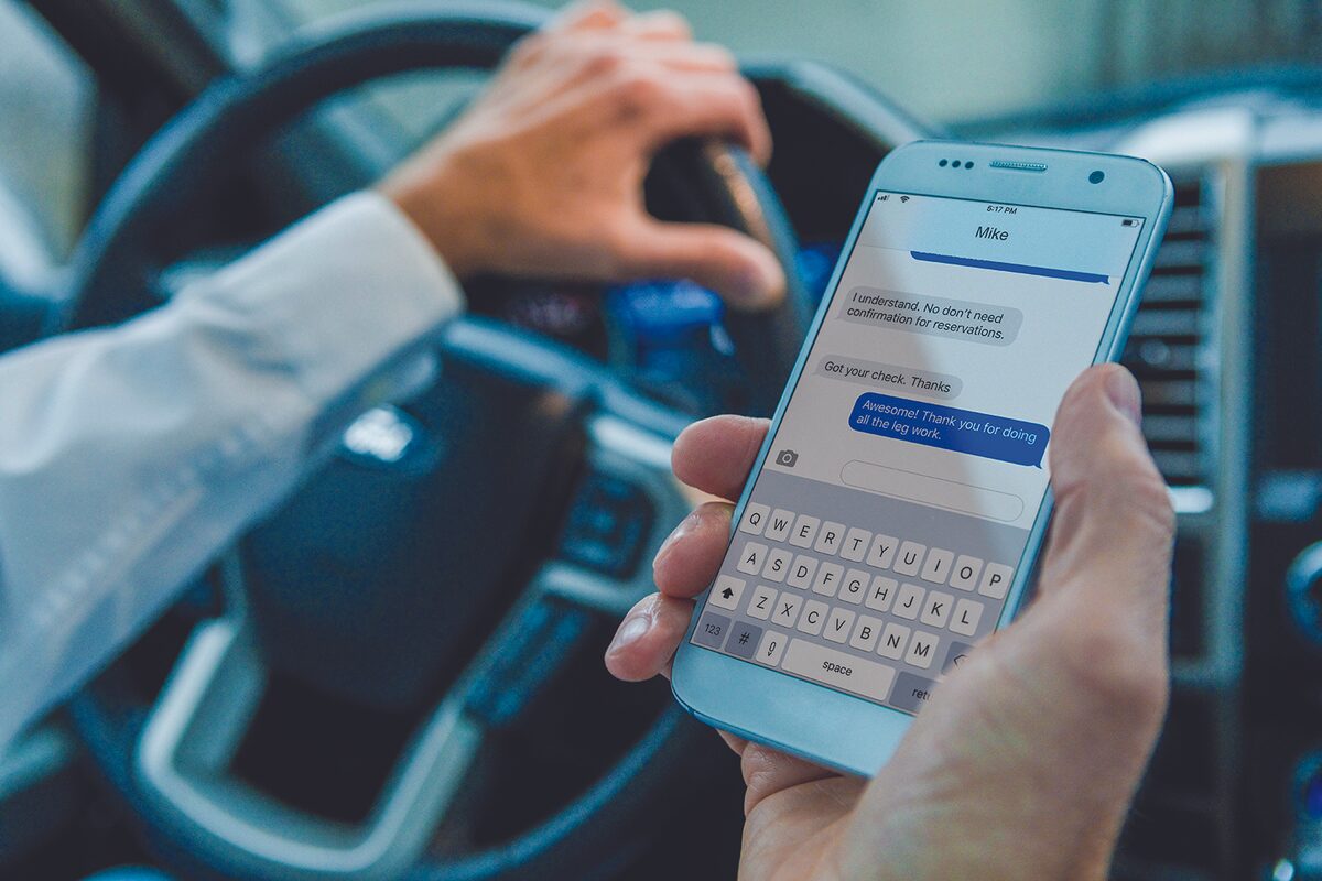 man holds cell phone and steering wheel while driving