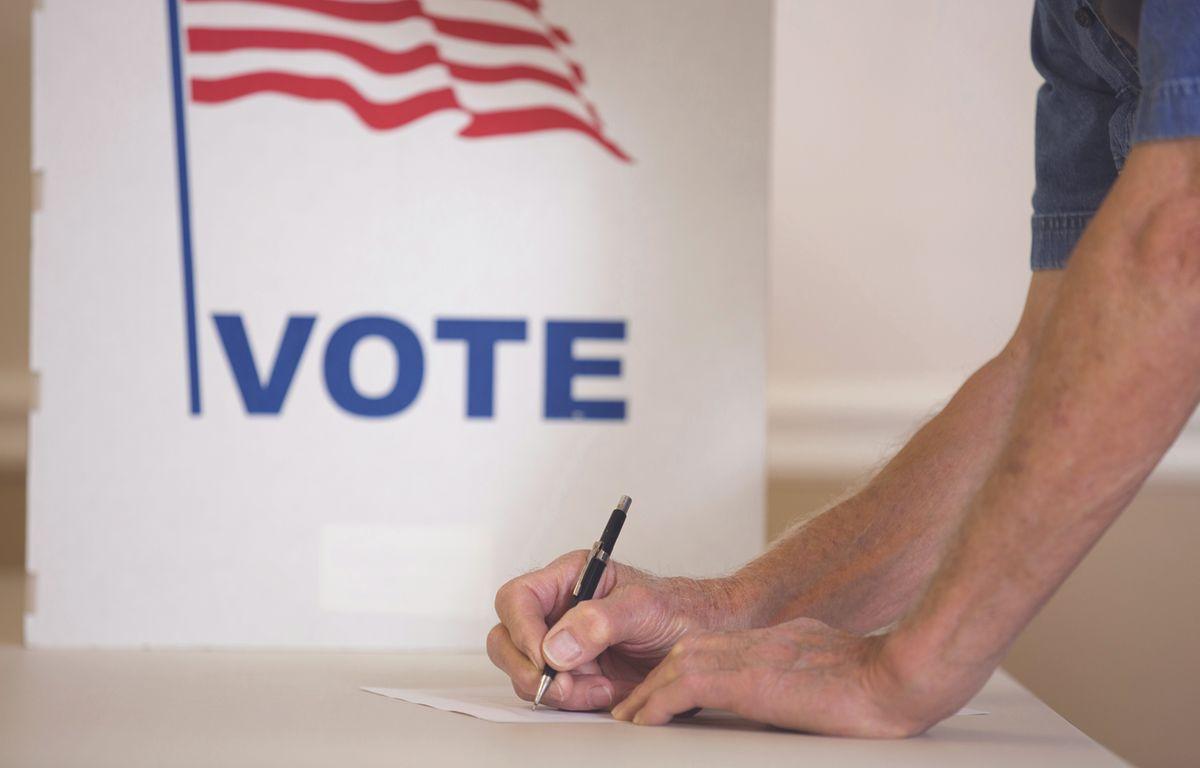 a man holds a pen to paper in front of a white sign that says vote with an american flag on it