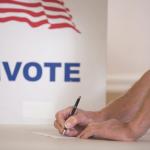 a man holds a pen to paper in front of a white sign that says vote with an american flag on it