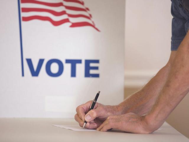 a man holds a pen to paper in front of a white sign that says vote with an american flag on it