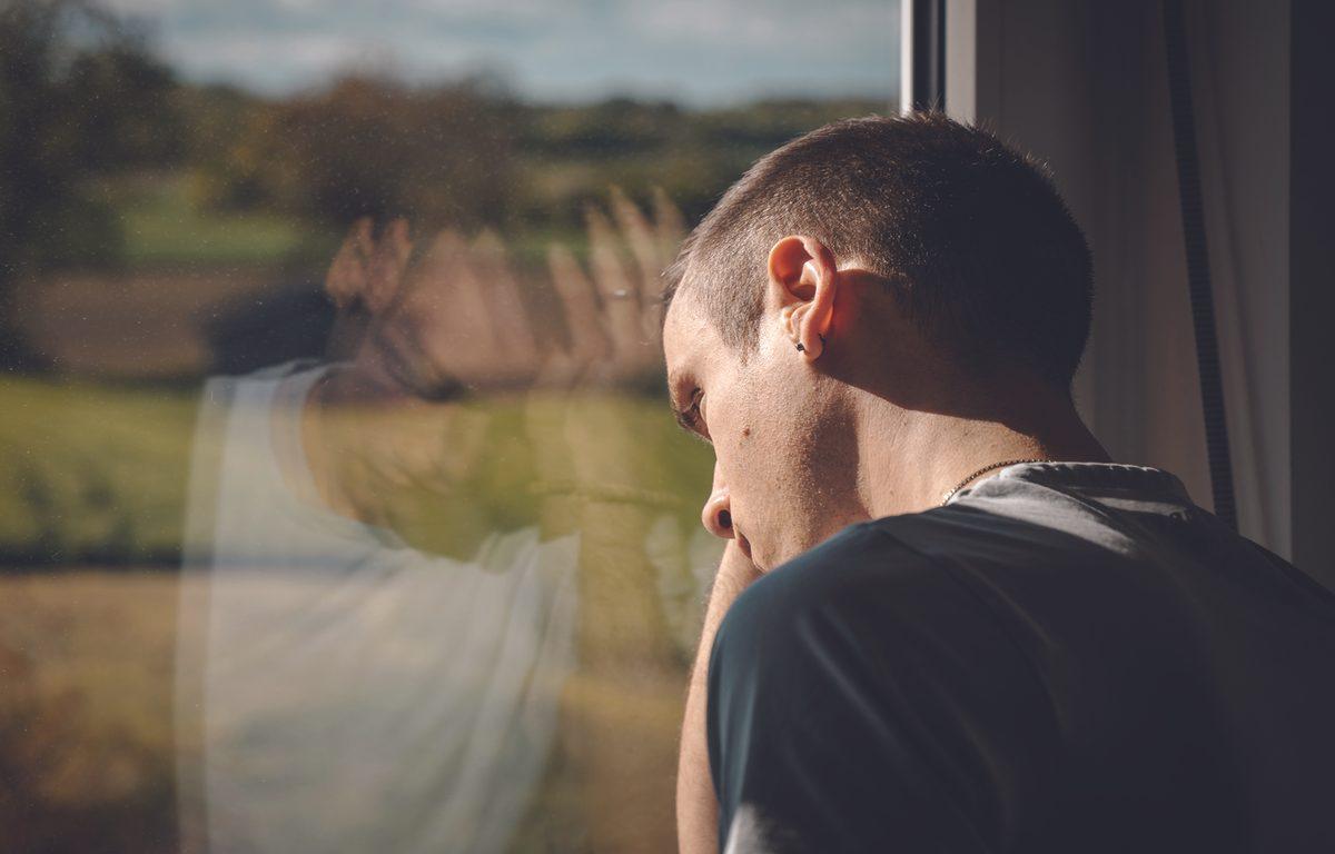 young man looks out window with hand on head