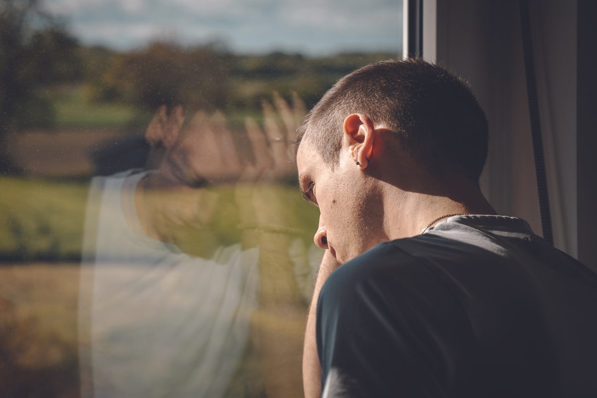 young man looks out window with hand on head