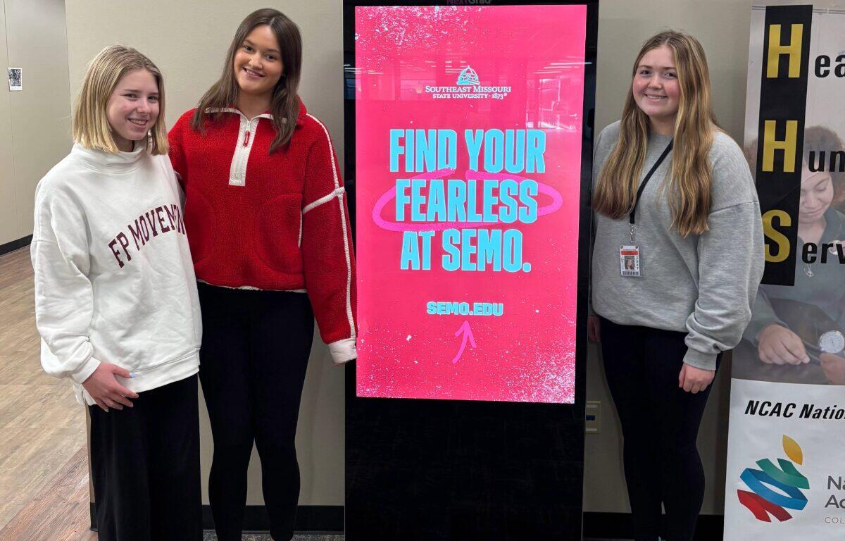 Three students stand around kiosk presenting colleges