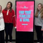 Three students stand around kiosk presenting colleges