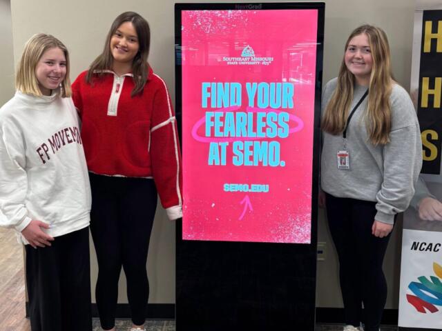 Three students stand around kiosk presenting colleges