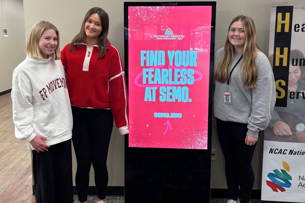Three students stand around kiosk presenting colleges