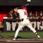 Baseball batter in a white uniform with number 10 swings at a pitch near home plate, catcher crouched behind him.
