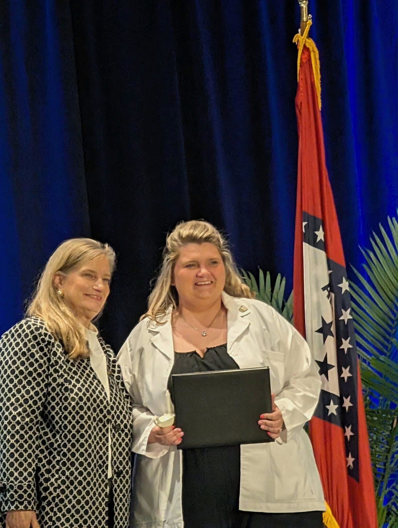 Two women stand on a stage during a ceremony; one in a white lab coat holds a black folder, smiling, with a flag in the background.