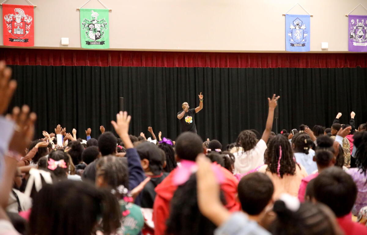 Male presenter on stage addressing a large crowd with hands raised in an auditorium settingy?