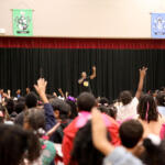Male presenter on stage addressing a large crowd with hands raised in an auditorium settingy?