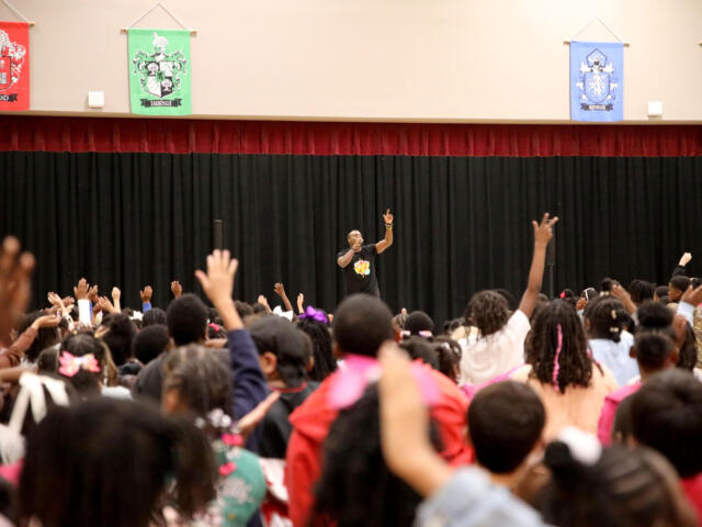 Male presenter on stage addressing a large crowd with hands raised in an auditorium settingy?
