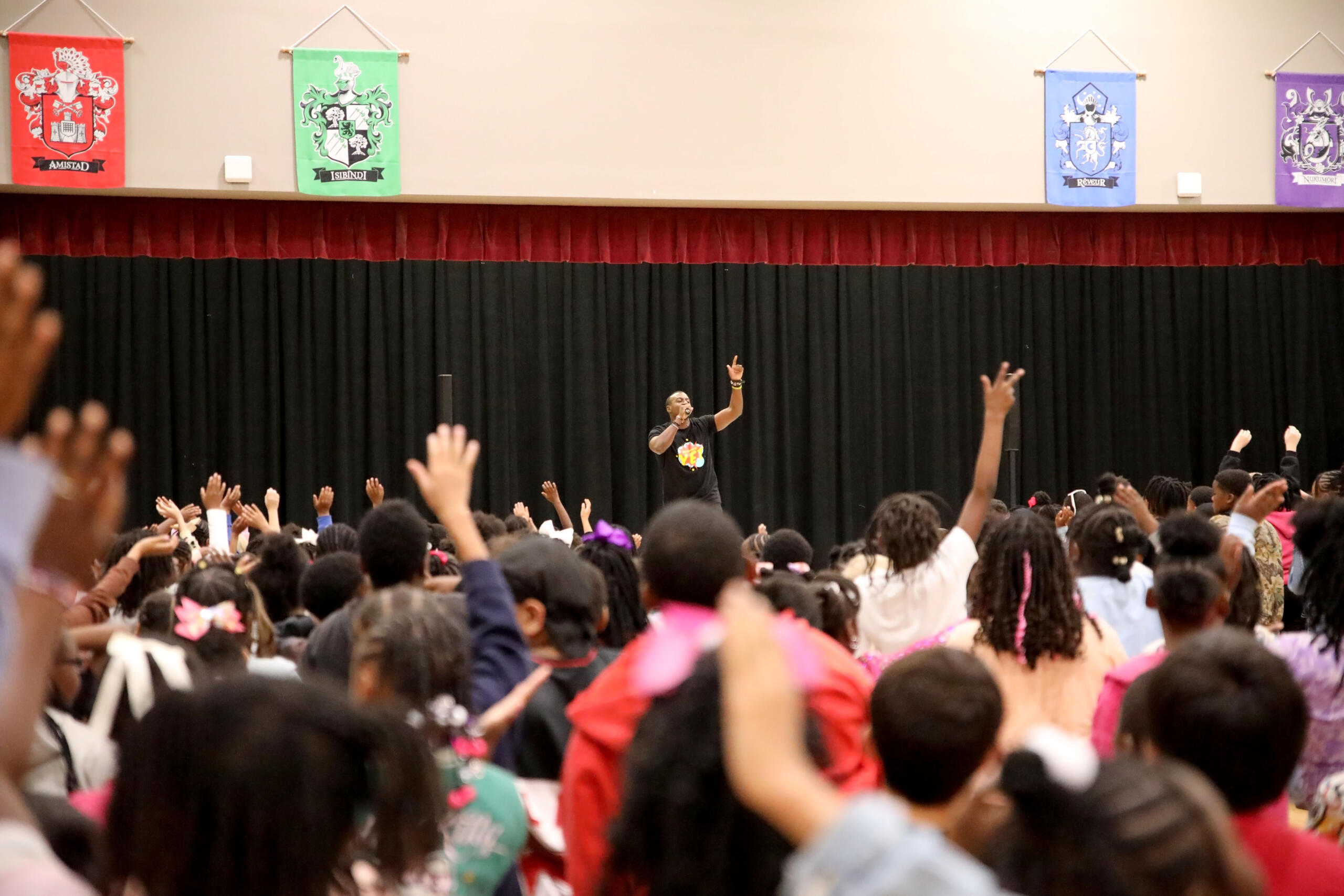 Male presenter on stage addressing a large crowd with hands raised in an auditorium settingy?