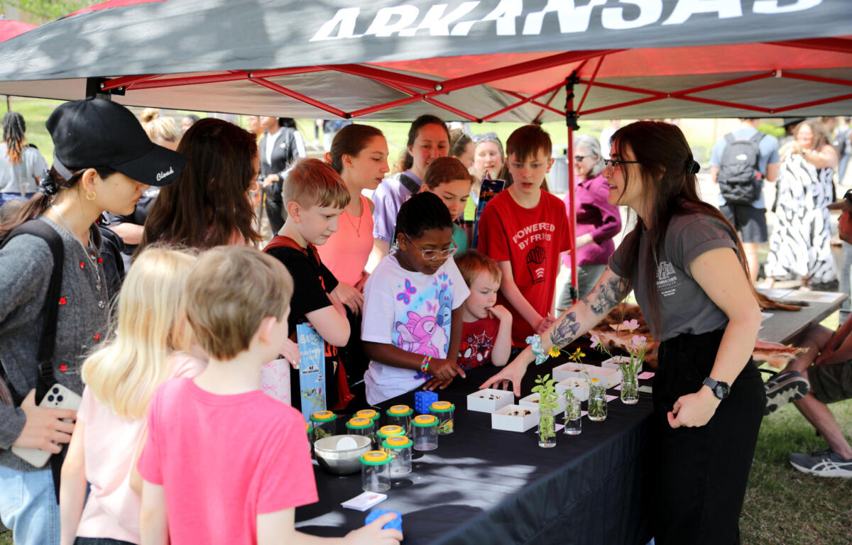 Children gather around a long table under a red-and-gray canopy, watching a woman with tattoos explain a craft with small plant jars and supplies on the table.