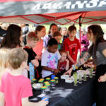 Children gather around a long table under a red-and-gray canopy, watching a woman with tattoos explain a craft with small plant jars and supplies on the table.