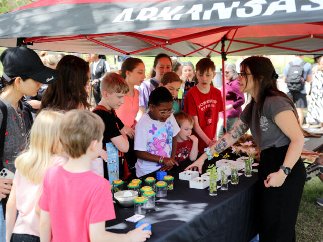 Children gather around a long table under a red-and-gray canopy, watching a woman with tattoos explain a craft with small plant jars and supplies on the table.
