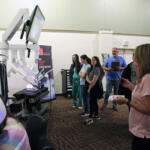 Group of people watching a row of white robotic arms on display in a science/tech booth.