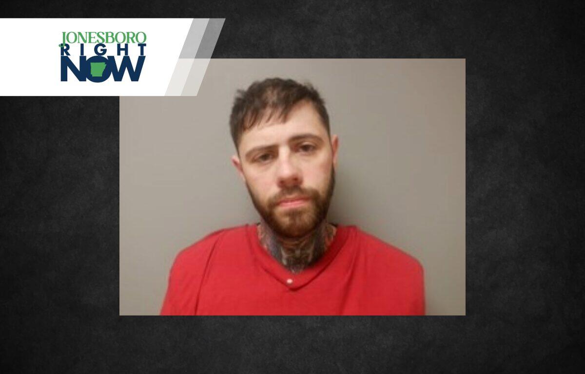 Mugshot-style photo of a man with a beard in a red shirt, facing the camera against a gray wall; a Jonesboro Right Now logo/banner overlaps the top-left corner.