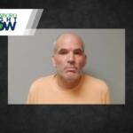 Mugshot of a middle-aged man with gray beard and short hair wearing an orange shirt against a gray backdrop.