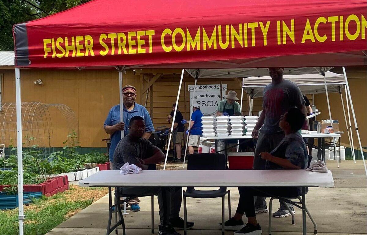 Volunteers under a red tent with 'Fisher Street Community in Action' banner, seated at a table with food trays in the background.