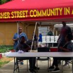 Volunteers under a red tent with 'Fisher Street Community in Action' banner, seated at a table with food trays in the background.