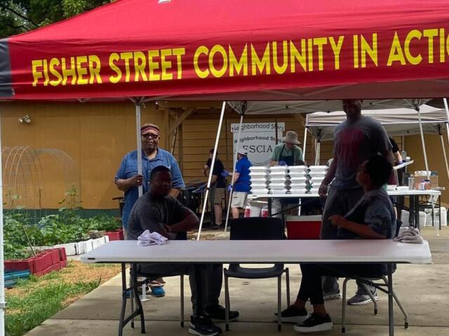 Volunteers under a red tent with 'Fisher Street Community in Action' banner, seated at a table with food trays in the background.