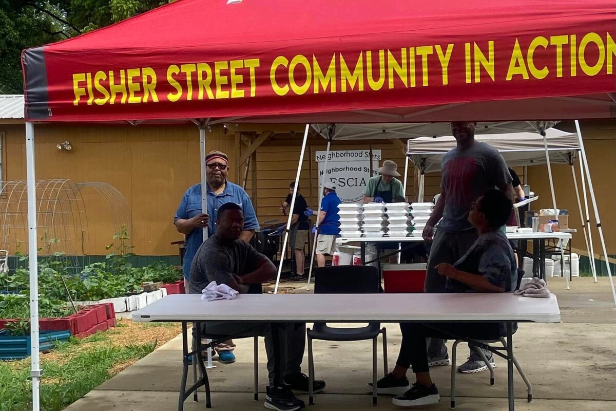 Volunteers under a red tent with 'Fisher Street Community in Action' banner, seated at a table with food trays in the background.