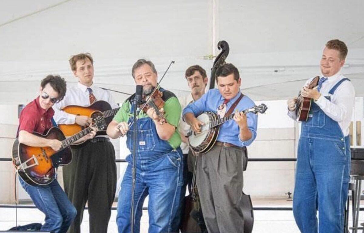 Six musicians perform a bluegrass-style set, playing guitars with a microphone at center stage.