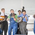 Six musicians perform a bluegrass-style set, playing guitars with a microphone at center stage.