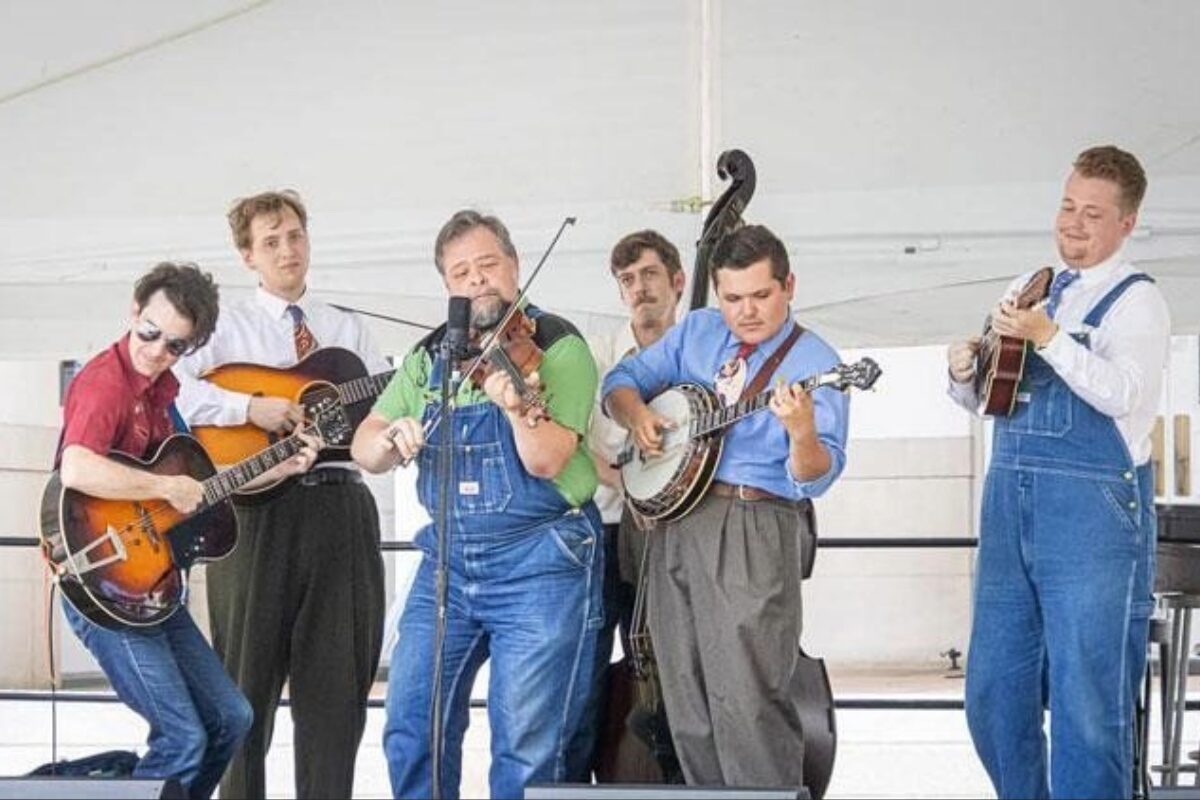 Six musicians perform a bluegrass-style set, playing guitars with a microphone at center stage.