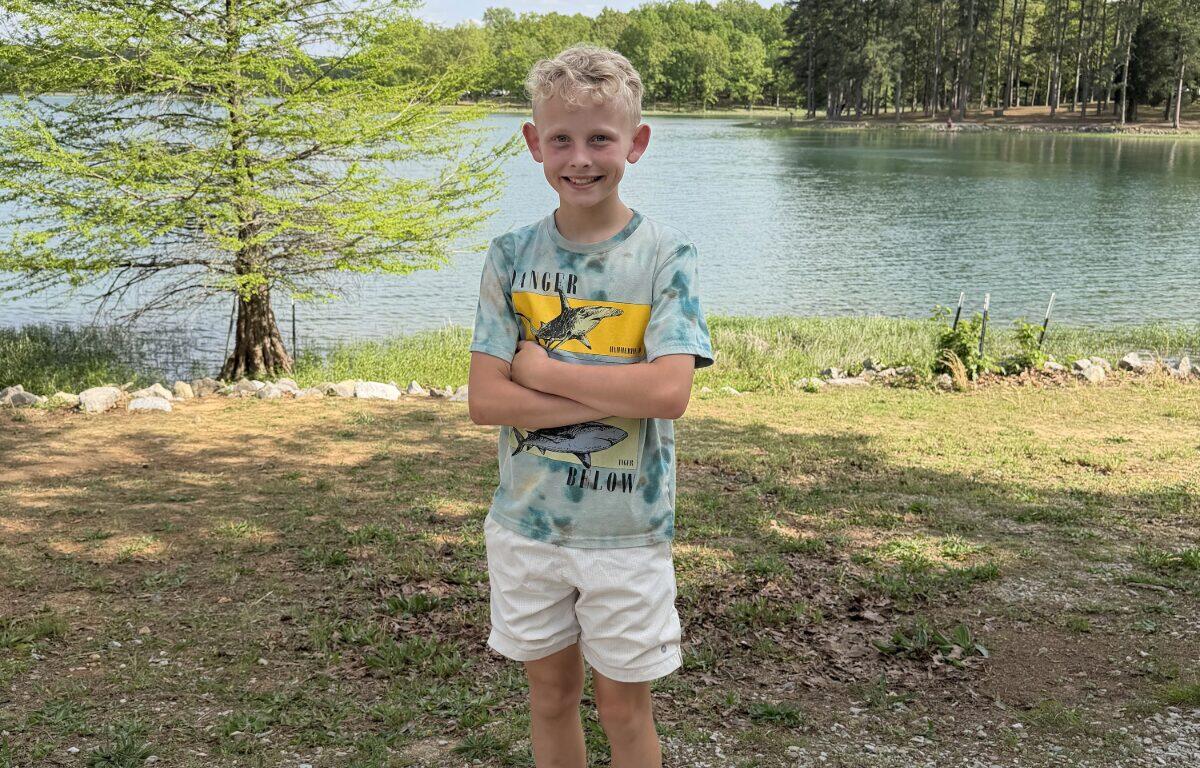 Smiling boy standing with arms crossed by a lakeside park, trees and water in the background