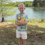 Smiling boy standing with arms crossed by a lakeside park, trees and water in the background