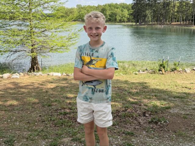 Smiling boy standing with arms crossed by a lakeside park, trees and water in the background
