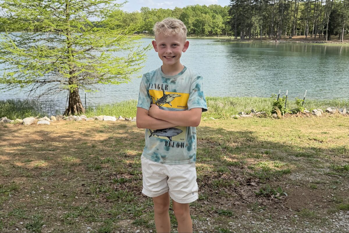 Smiling boy standing with arms crossed by a lakeside park, trees and water in the background