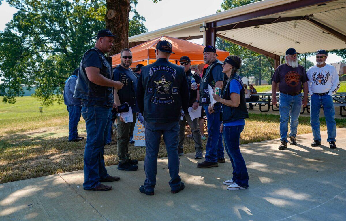 Group of bikers in leather vests and patches stand in a semicircle outdoors, chatting near a pavilion at a park event.