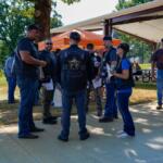 Group of bikers in leather vests and patches stand in a semicircle outdoors, chatting near a pavilion at a park event.