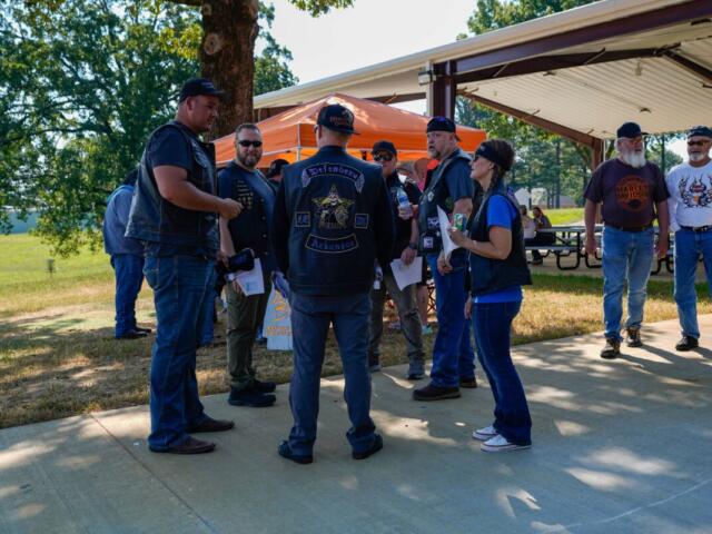 Group of bikers in leather vests and patches stand in a semicircle outdoors, chatting near a pavilion at a park event.