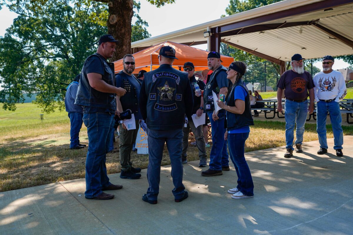 Group of bikers in leather vests and patches stand in a semicircle outdoors, chatting near a pavilion at a park event.