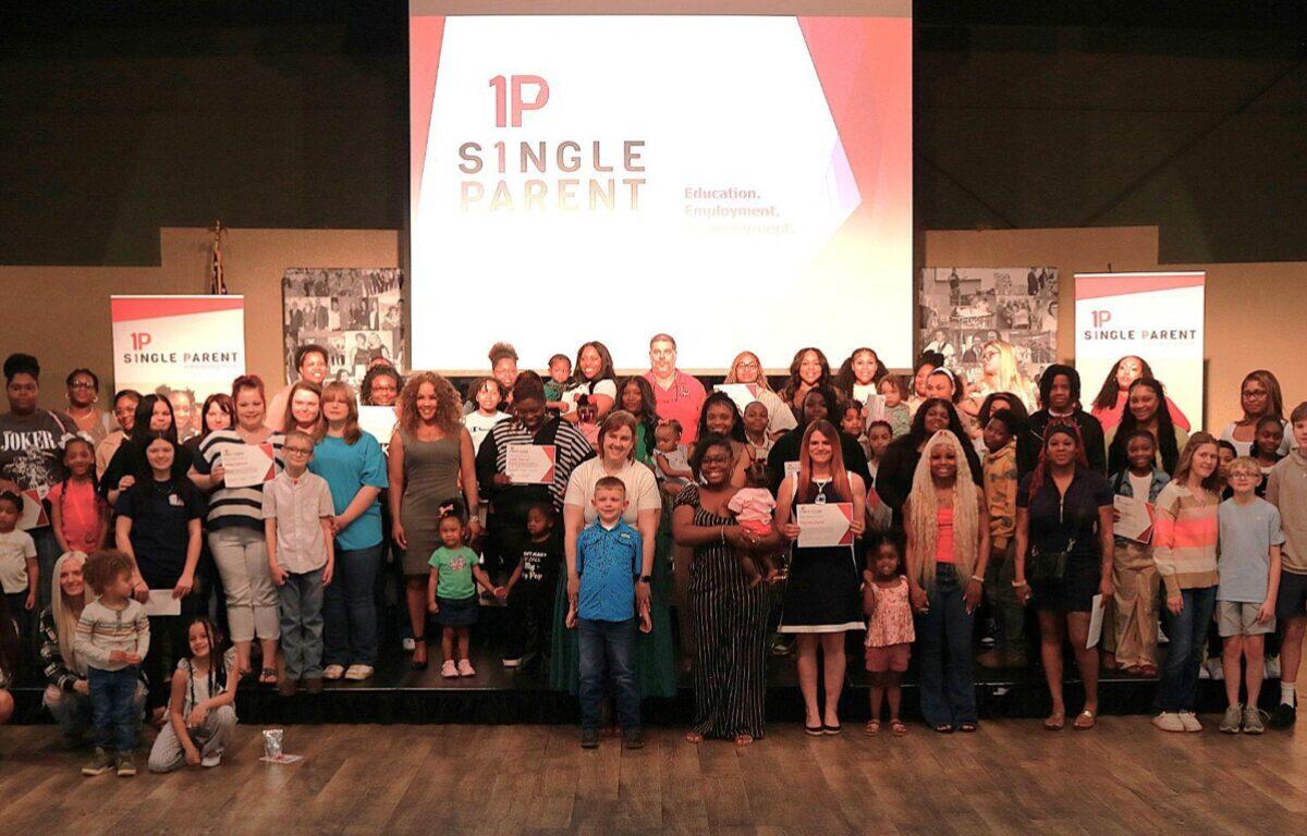 Group of diverse adults and children on a stage, many holding certificates, at a 1P Single Parent event with a projector logo backdrop.