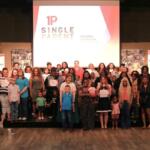 Group of diverse adults and children on a stage, many holding certificates, at a 1P Single Parent event with a projector logo backdrop.