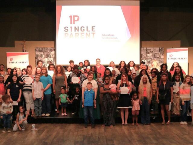 Group of diverse adults and children on a stage, many holding certificates, at a 1P Single Parent event with a projector logo backdrop.