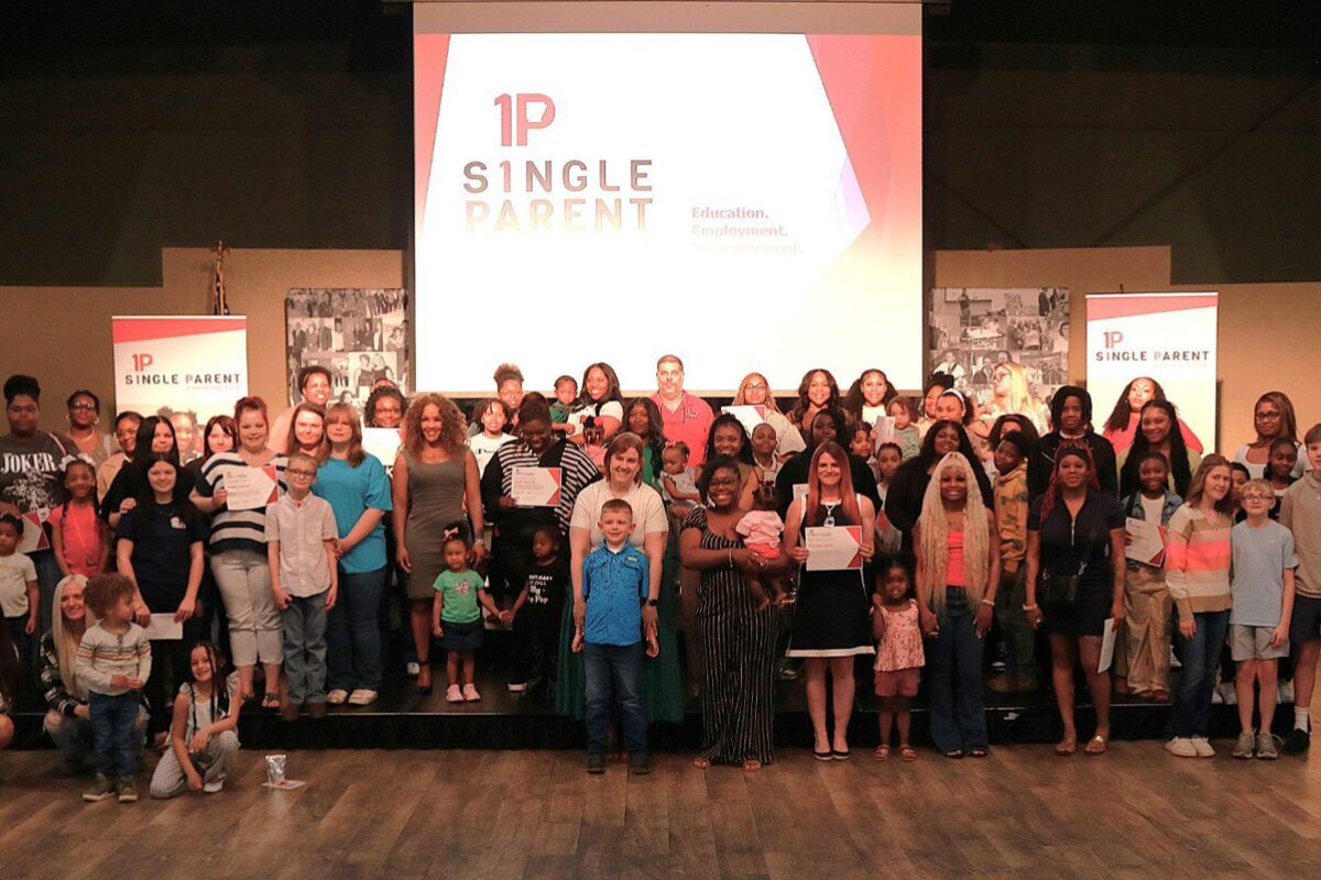 Group of diverse adults and children on a stage, many holding certificates, at a 1P Single Parent event with a projector logo backdrop.
