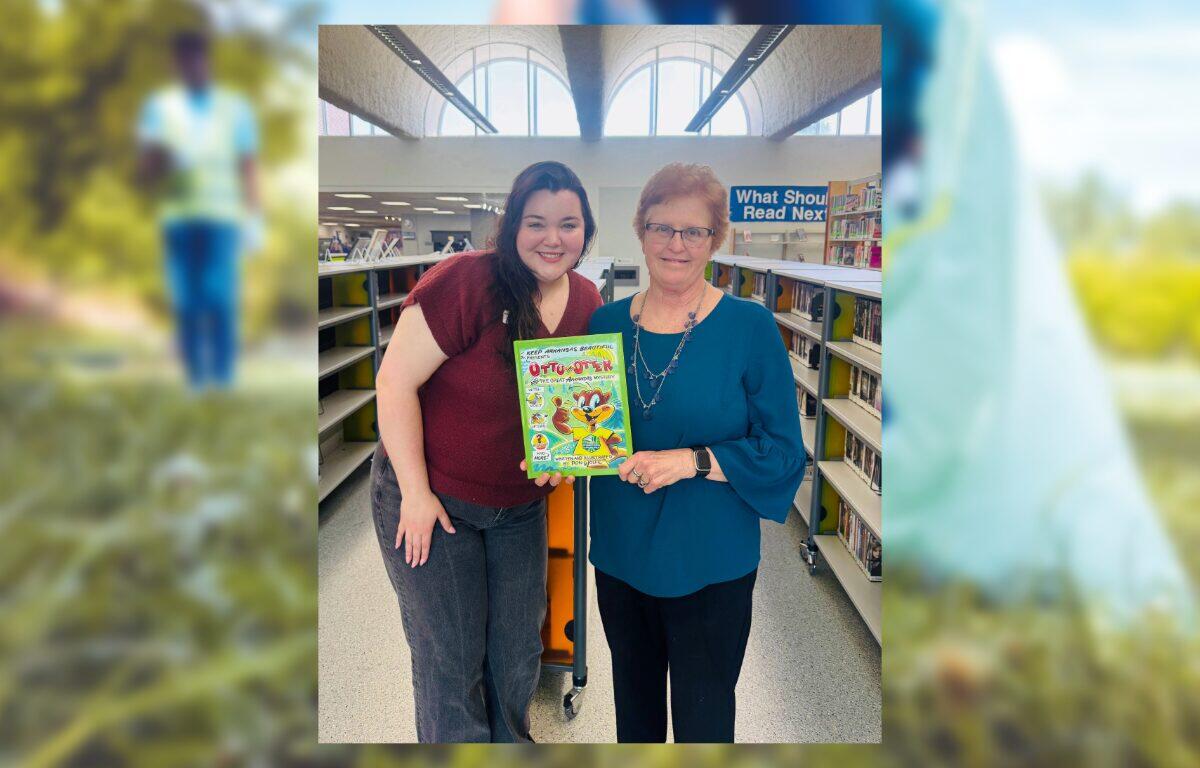 Two women in a library pose together, smiling, holding a bright cartoon book between them.