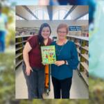 Two women in a library pose together, smiling, holding a bright cartoon book between them.