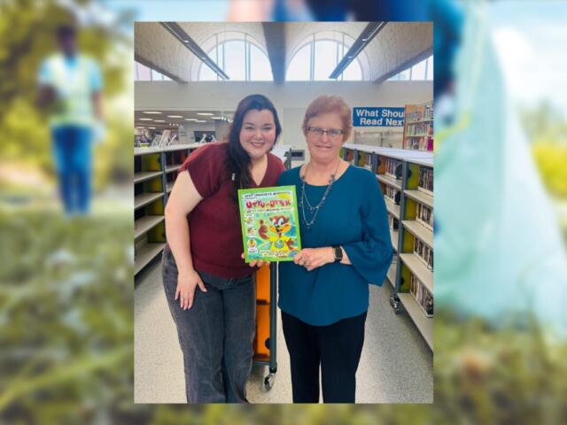 Two women in a library pose together, smiling, holding a bright cartoon book between them.