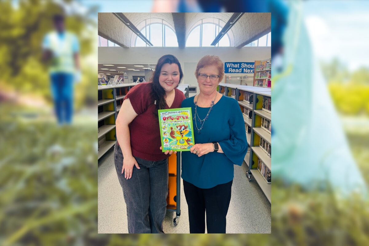 Two women in a library pose together, smiling, holding a bright cartoon book between them.