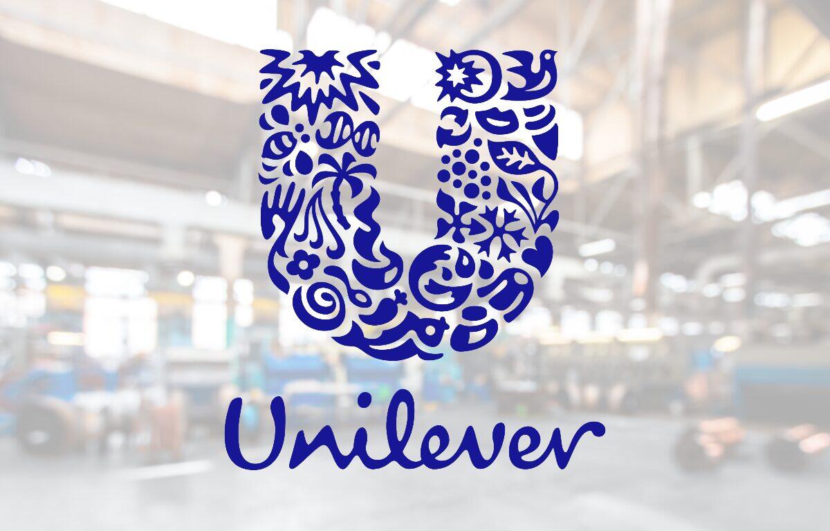 Blue Unilever logo: a decorative U made of floral shapes with the word Unilever beneath, shown on a glass surface in a blurred industrial background.