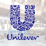 Blue Unilever logo: a decorative U made of floral shapes with the word Unilever beneath, shown on a glass surface in a blurred industrial background.