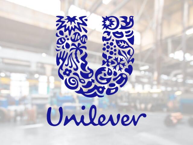 Blue Unilever logo: a decorative U made of floral shapes with the word Unilever beneath, shown on a glass surface in a blurred industrial background.